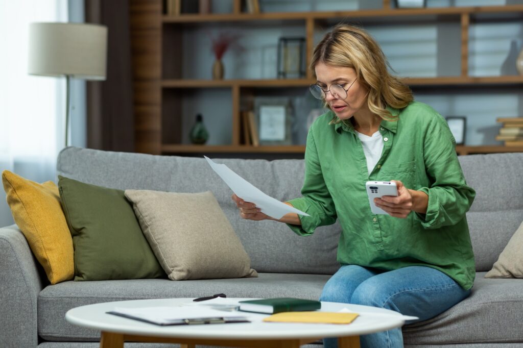 Senior woman of retirement age sitting at home on sofa with phone and checking documents, bank